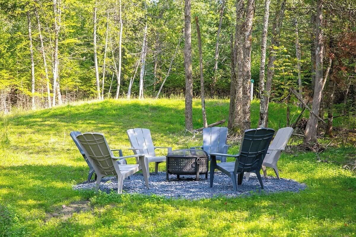A group of Adirondack chairs arranged around a metal fire pit in a grassy outdoor area surrounded by trees.