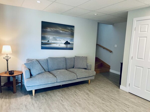 A living room with a grey sofa, a small round table with a lamp, and a painting of a seascape on the wall. - Edge of Lake Lovering - Spa - Crépuscule (Memphrémagog)