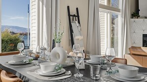 Une table à manger dressée avec de l’assiette blanche, des bols et des verres à vin, avec une vue sur la mer et les montagnes en arrière-plan.