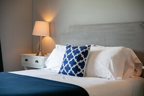 A neatly made bed with white and blue pillows, a blue throw blanket, and a bedside lamp on a white nightstand.