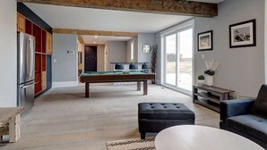 A spacious living area with a pool table, a modern kitchen with a refrigerator, and a large window providing a view of the outdoors.