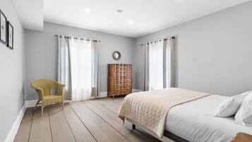Een slaapkamer met een bed, een houten dressoir, een rieten stoel en een spiegel aan de muur.