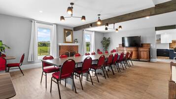 A spacious dining area with a long table and red chairs, a fireplace, and a kitchen area with cabinets and a TV.