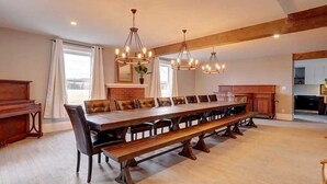 A spacious dining area with a long wooden table and leather chairs, a piano, and a kitchen in the background.