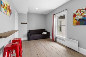 Una habitación con un sofá oscuro, suelo de madera, una silla roja, un radiador blanco y una ventana con cortinas rojas.