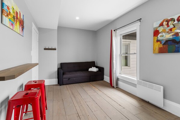 A room with a dark sofa, a wooden floor, a red chair, a white radiator, and a window with red curtains.