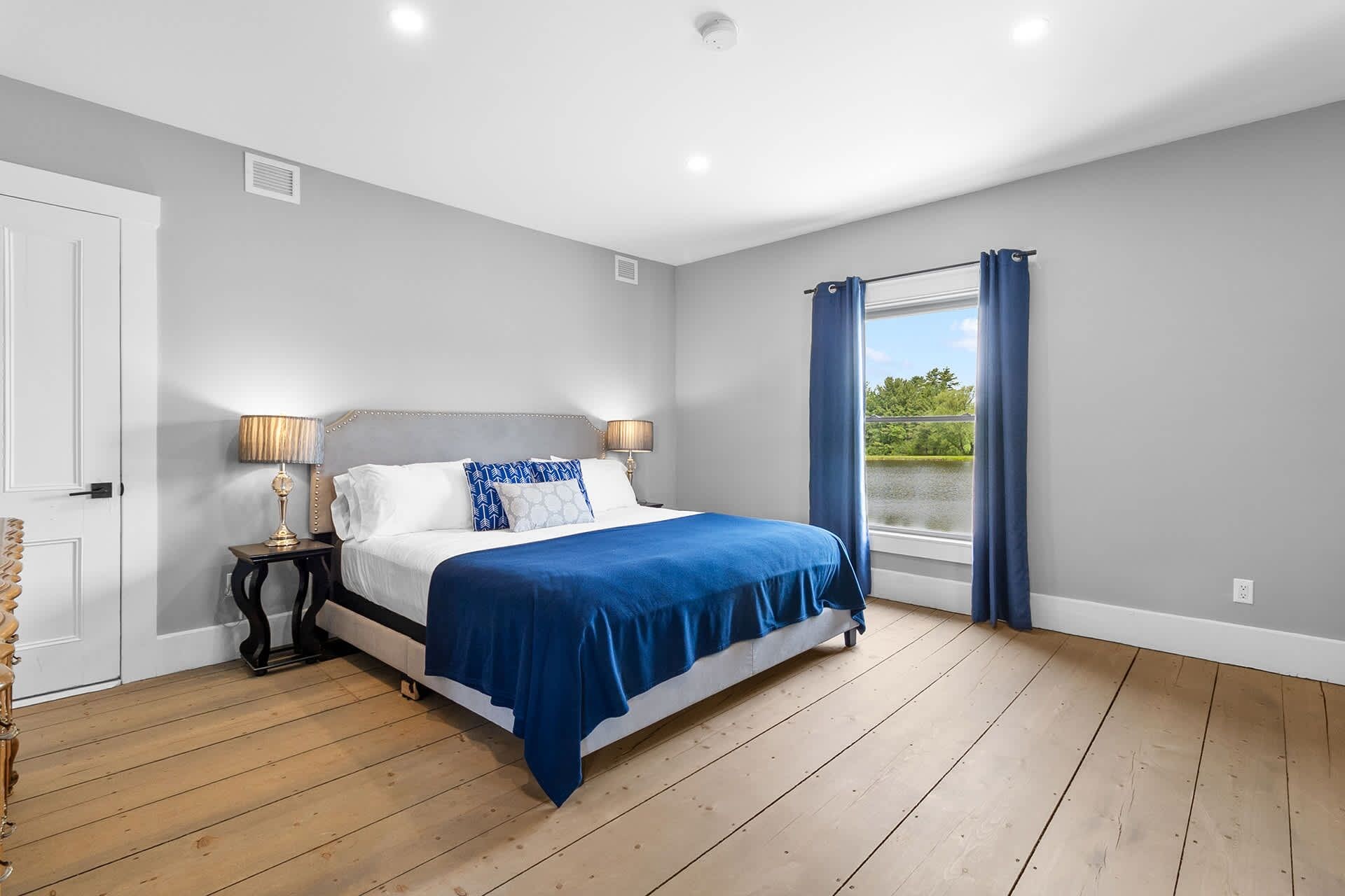 A bedroom with a large bed, a nightstand, a lamp, a window with curtains, and a view of greenery outside.