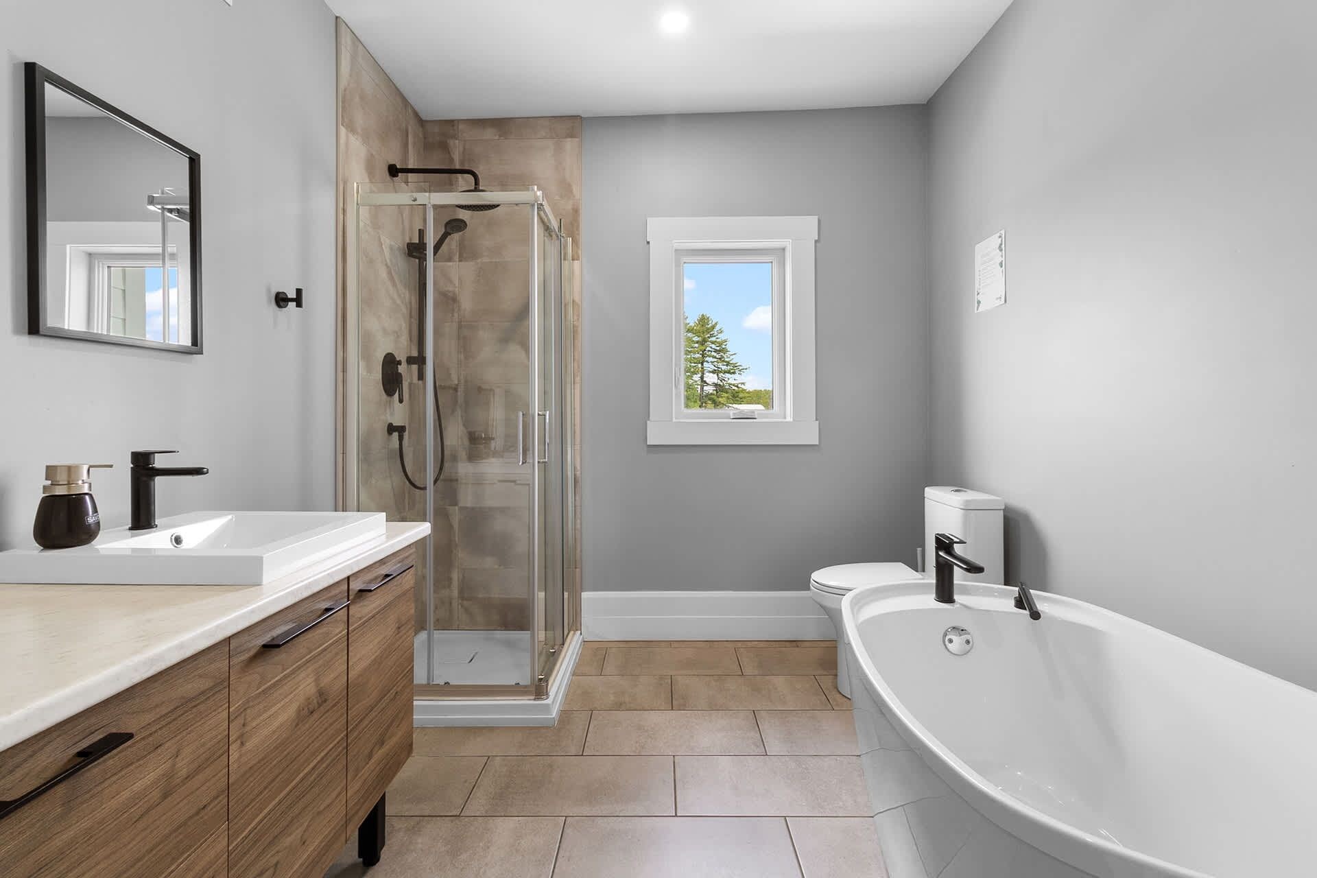 Un baño moderno con ducha envuelta en vidrio, una bañera grande y una ventana con vista a árboles.