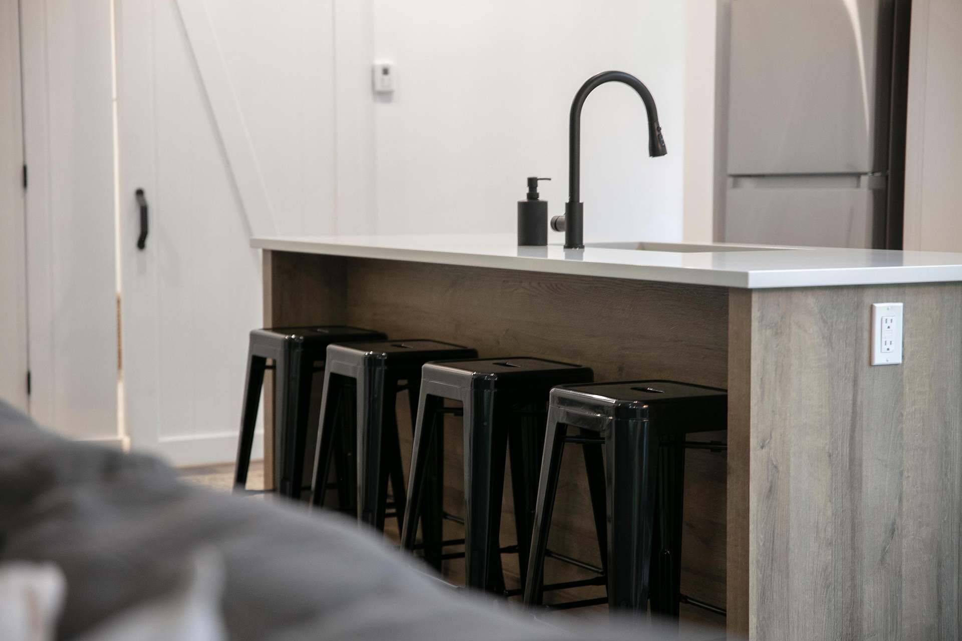 Eine kleine Küche mit weißer Arbeitsplatte, schwarzen Barhocker und einem schwarzen Wasserhahn.