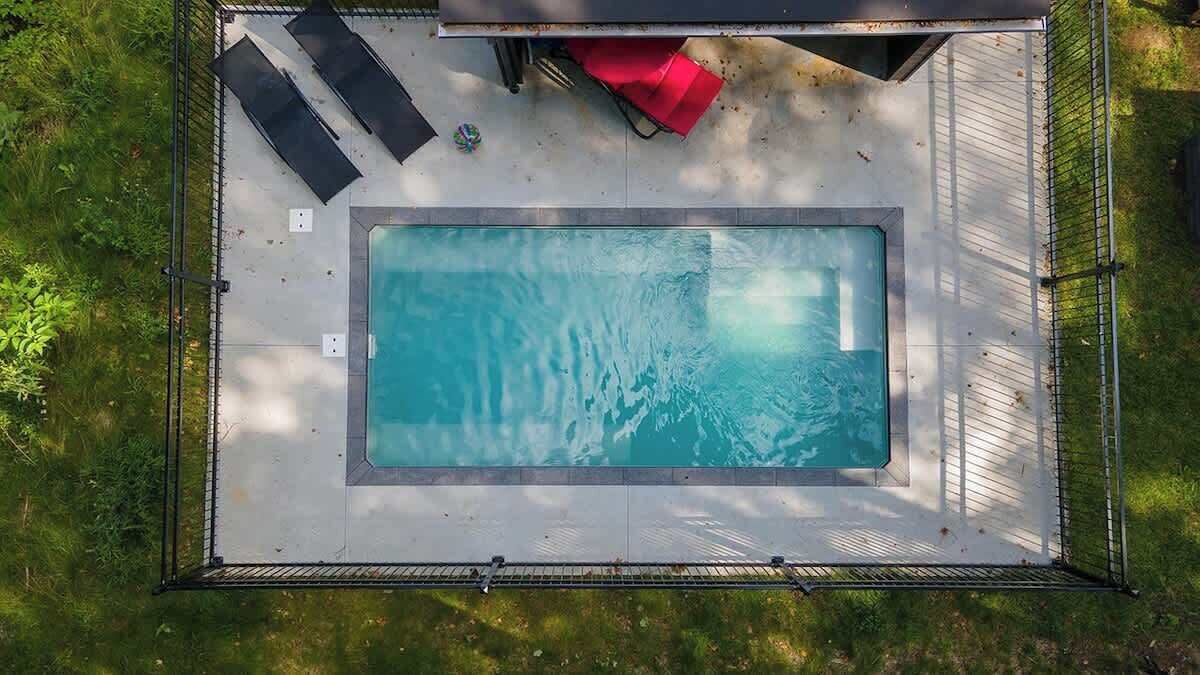 An aerial view of a rectangular swimming pool with a surrounding deck, a metal fence, and a small red object on the deck.