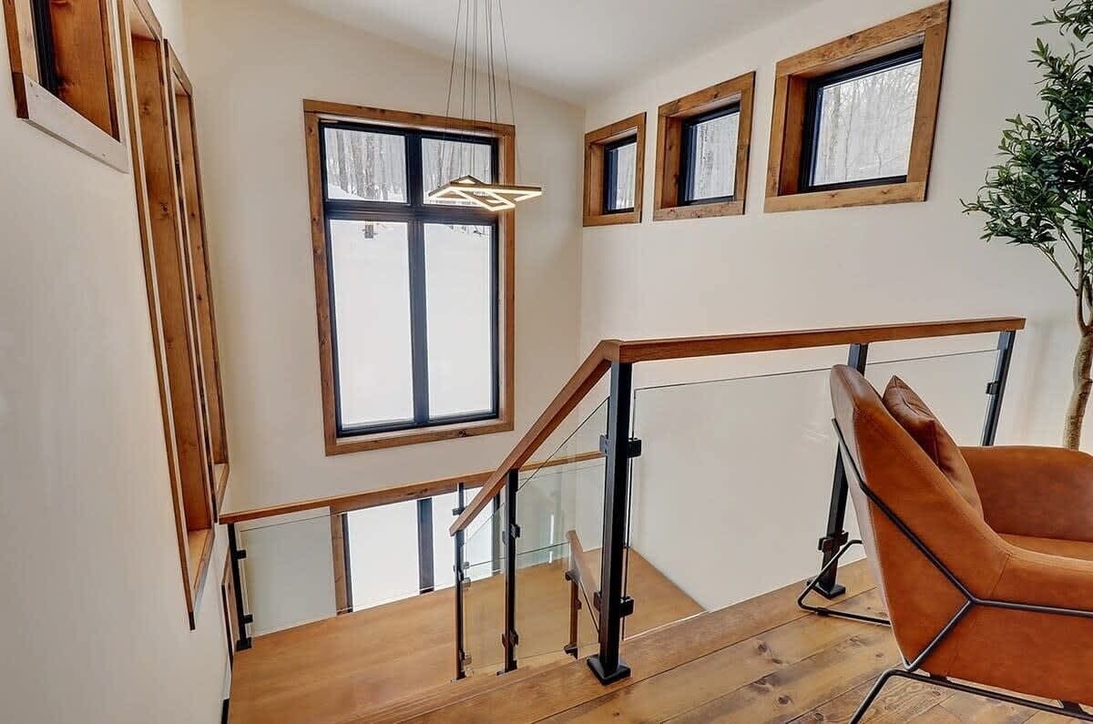 Una moderna scala con ringhiere in legno e parapetti in vetro, che conduce a un secondo piano con ampie finestre e un lampadario.