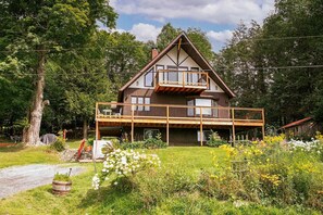 A two-story wooden house with a balcony, surrounded by greenery and a clear sky.