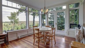 A dining area with a round table and wooden chairs, large windows with a view of trees, and a chandelier. - Brome Lake - Inground Pool - Spa - Lumen (Knowlton)