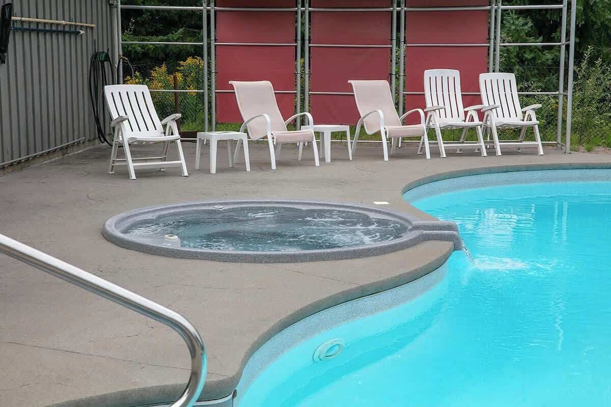 A swimming pool with a hot tub, surrounded by white lounge chairs and a red wall in the background.