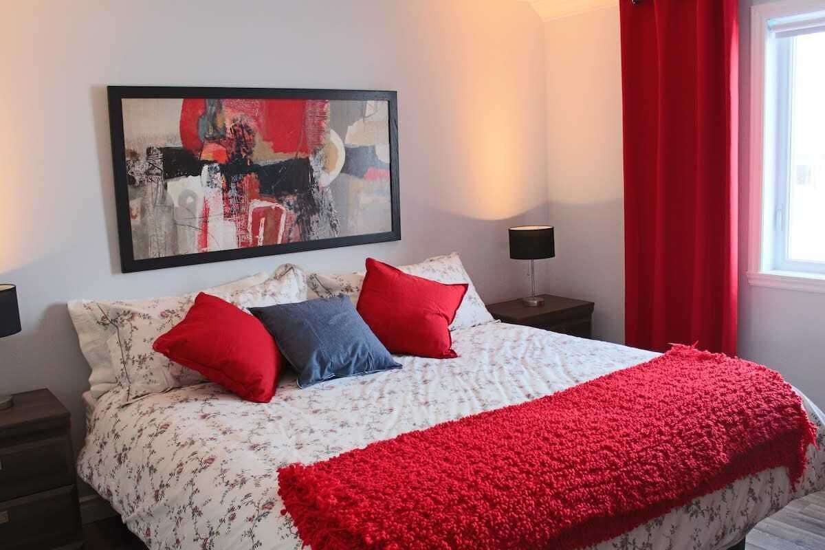 A neatly made bed with floral bedding and red accents, a modern abstract painting above it, and a window with red curtains.