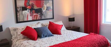 A neatly made bed with floral bedding and red accents, a modern abstract painting above it, and a window with red curtains.