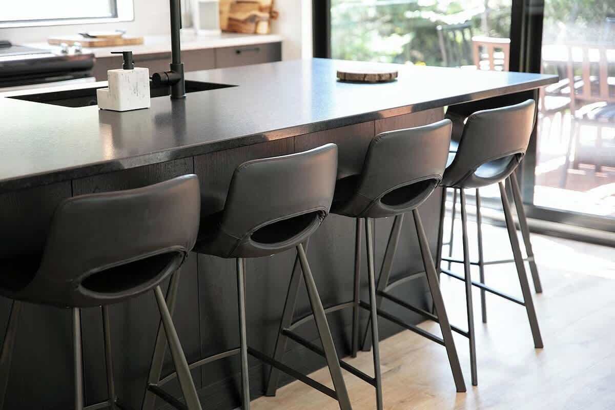 Une cuisine moderne avec un plan de travail foncé, quatre tabourets de bar et une fenêtre donnant sur l’extérieur.