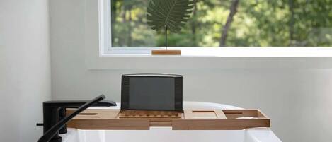 A white desk with a black monitor, wooden side table, and a leaf decoration on a window sill.