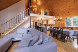 A spacious living area with a grey sectional sofa, a dining table with chairs, a staircase leading to an upper level, and a kitchen area.