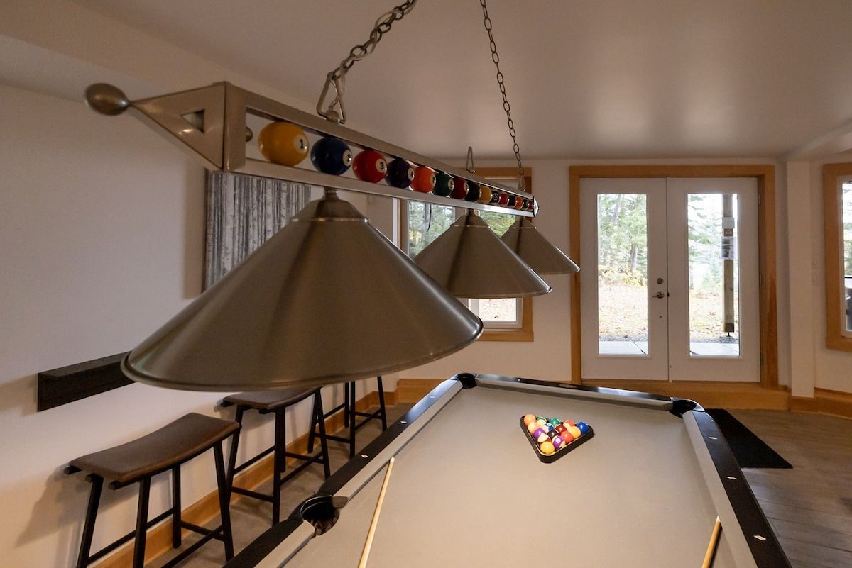 Una mesa de billar con un juego de bolas, iluminación colgante y una ventana al fondo.