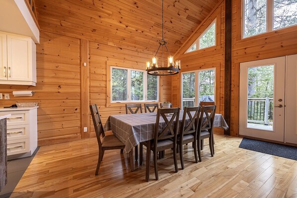 Un interior de madera con una mesa de comedor y sillas, una araña y ventanas amplias.