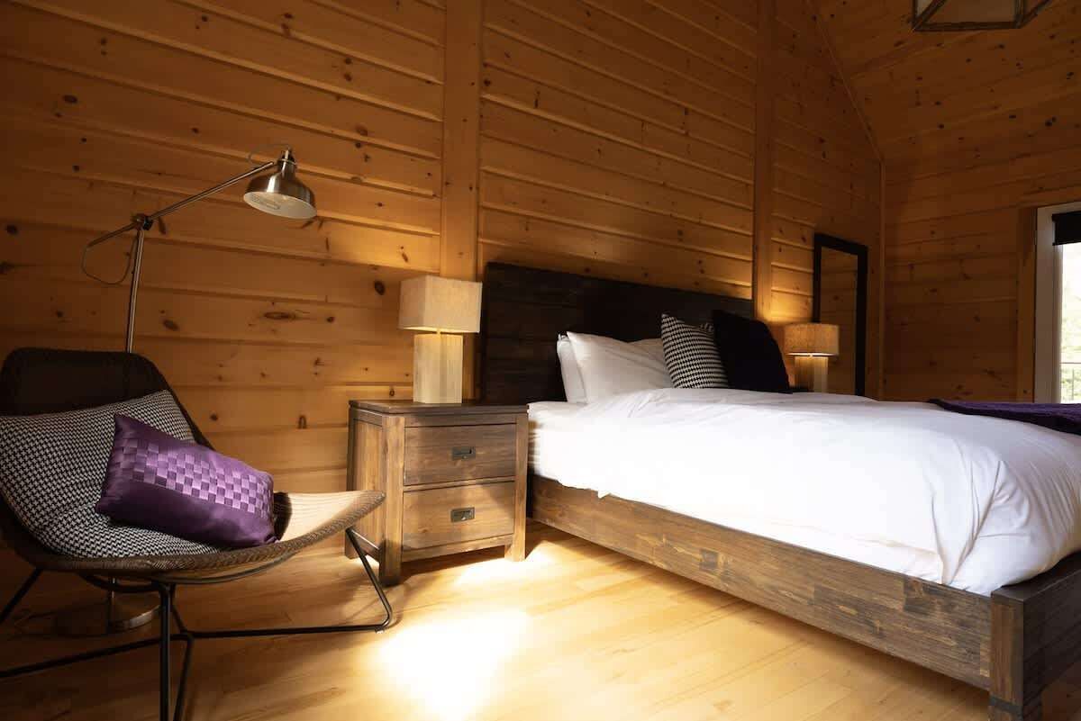 A wooden cabin bedroom with a bed, nightstands, a chair, and a lamp.