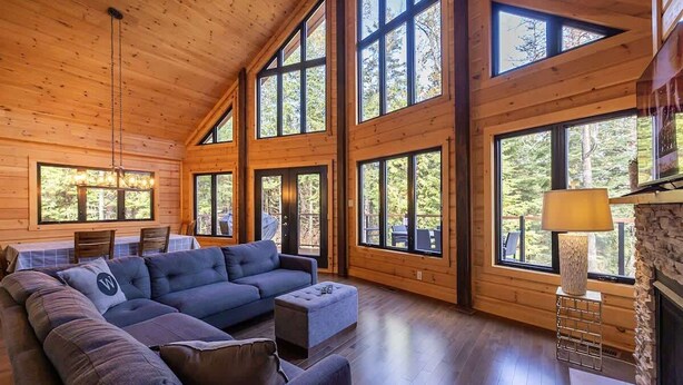 Un salon spacieux doté d’un grand canapé d’angle, d’une cheminée et de grandes fenêtres offrant une vue sur la forêt environnante.