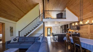 Un vaste espace de vie comprenant un canapé gris, une table à manger avec des chaises, une cuisine dotée de meubles en bois et un escalier menant à l’étage supérieur.