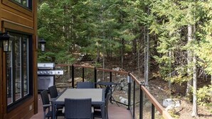 Une terrasse en bois avec une table et des chaises, entourée d’une rambarde en verre, donnant sur une zone boisée.