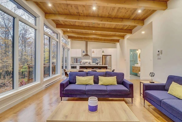Un intérieur moderne avec un plafond en bois, de grandes fenêtres, un canapé violet et une cuisine à l’arrière-plan.