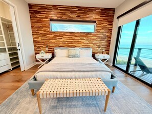A modern bedroom with a wooden wall, a large bed, bedside tables, a bench, and a view of the ocean.