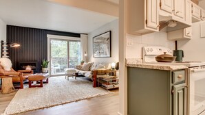 Una sala de estar moderna con chimenea, sofá y mesa de centro. Se observa una cocina con armarios blancos y una estufa.