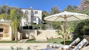 A pool area with lounge chairs and an umbrella, surrounded by a stone wall and lush greenery. - Charming villa with garden and heated pool, perfect families (Saint Paul de Vence)