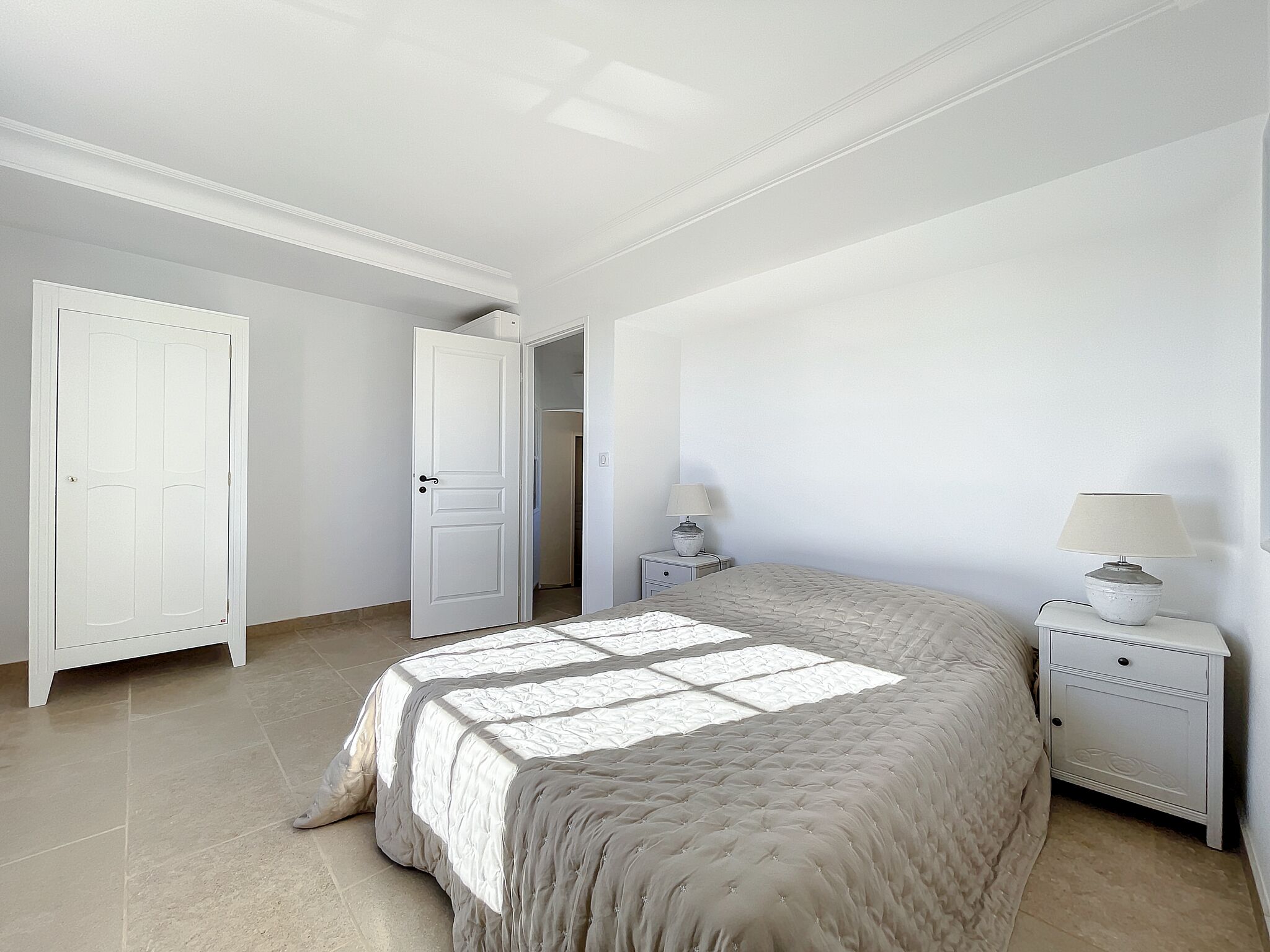 A bedroom with a large bed, a nightstand with a lamp, and a door leading to another room.