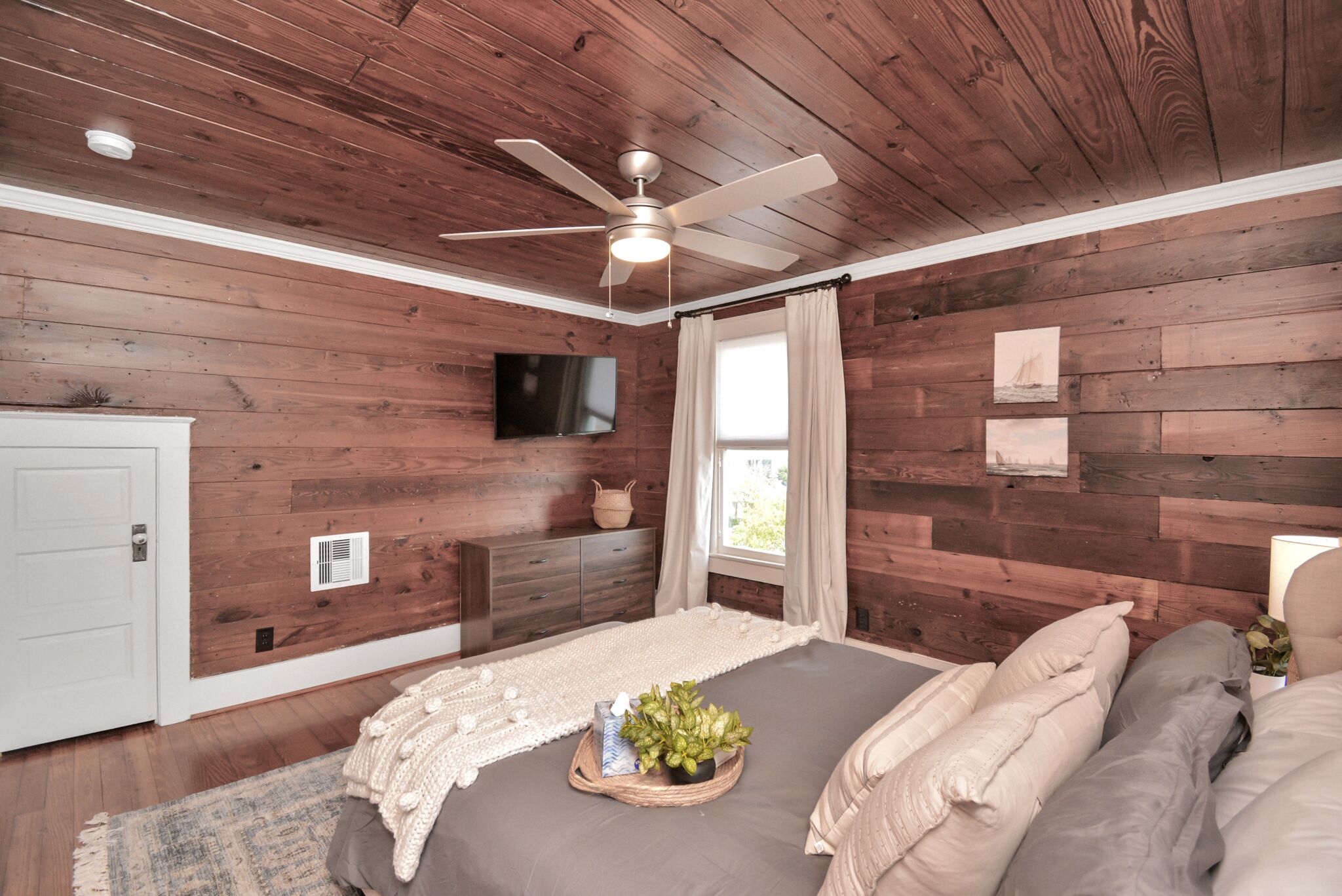 A bedroom with wooden walls, a ceiling fan, a bed with pillows, a bedside table with a lamp, and a window with curtains.