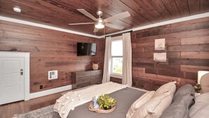 A bedroom with wooden walls, a ceiling fan, a bed with pillows, a bedside table with a lamp, and a window with curtains.