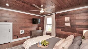 A bedroom with wooden walls, a ceiling fan, a bed with pillows, a bedside table with a lamp, and a window with curtains.