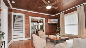 A spacious living room with a wooden floor, a ceiling fan, a flat-screen TV mounted on the wall, and a staircase leading to an upper level.