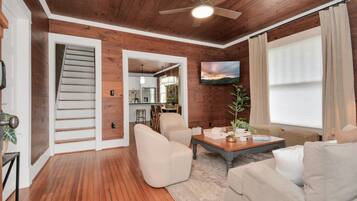A spacious living room with a wooden floor, a ceiling fan, a flat-screen TV mounted on the wall, and a staircase leading to an upper level.