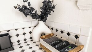 Un bagno con pavimento piastrellato di bianco, un vaso bianco con vegetazione scura e un cesto con asciugamani contenenti la scritta 'makeup'.