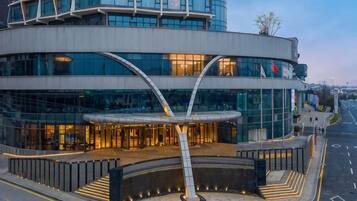 A modern building with a curved glass facade, a prominent entrance with a metal archway, and a well-lit exterior.