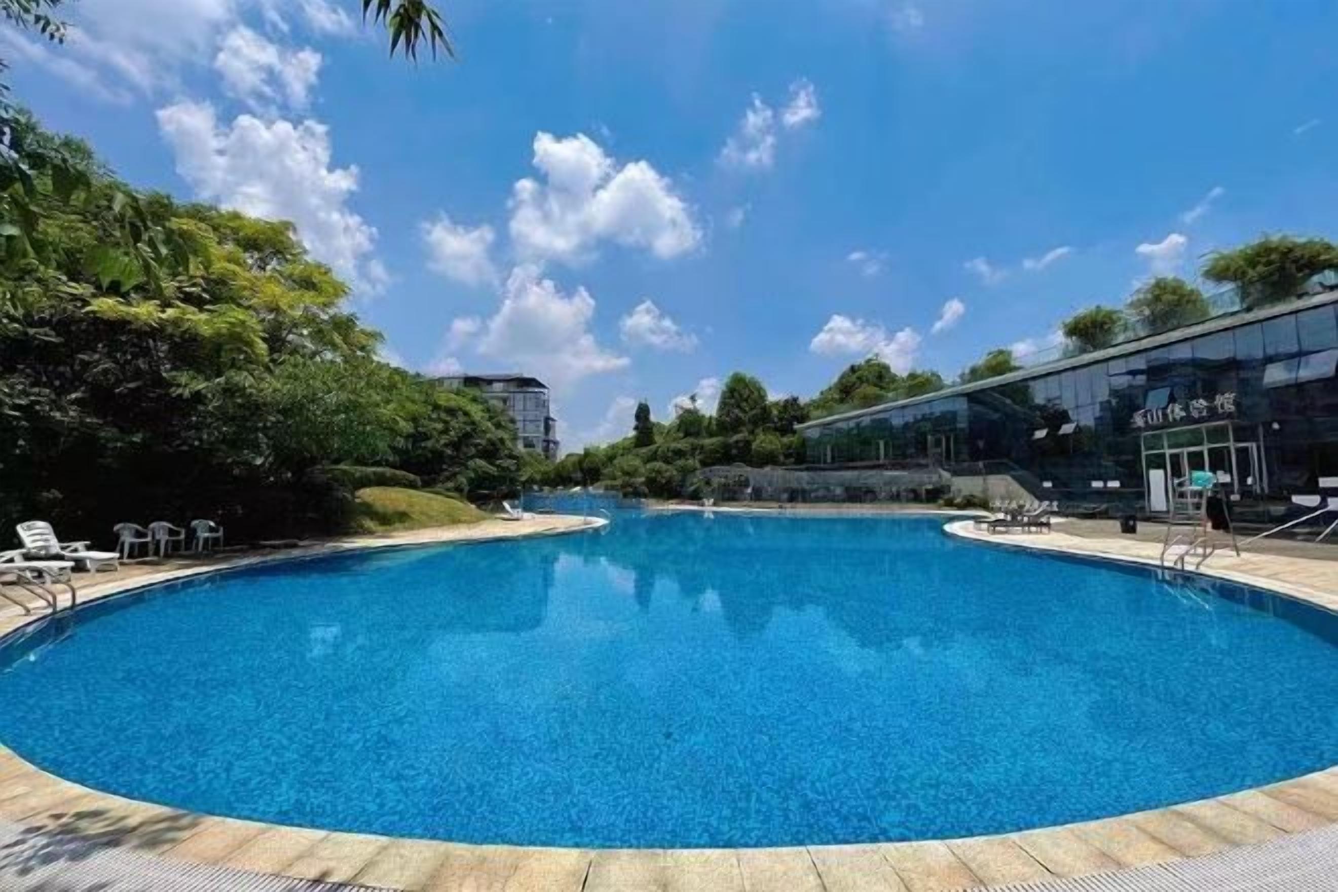 A large, clear swimming pool with lounge chairs and a modern building in the background.