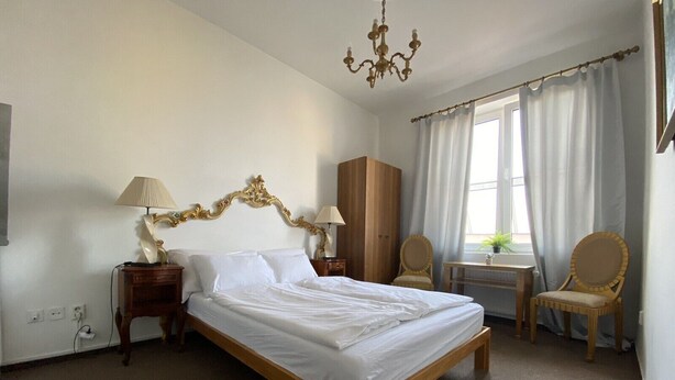 A hotel room with a large bed, two bedside tables, a chandelier, a wardrobe, a desk with a chair, and a window with curtains.