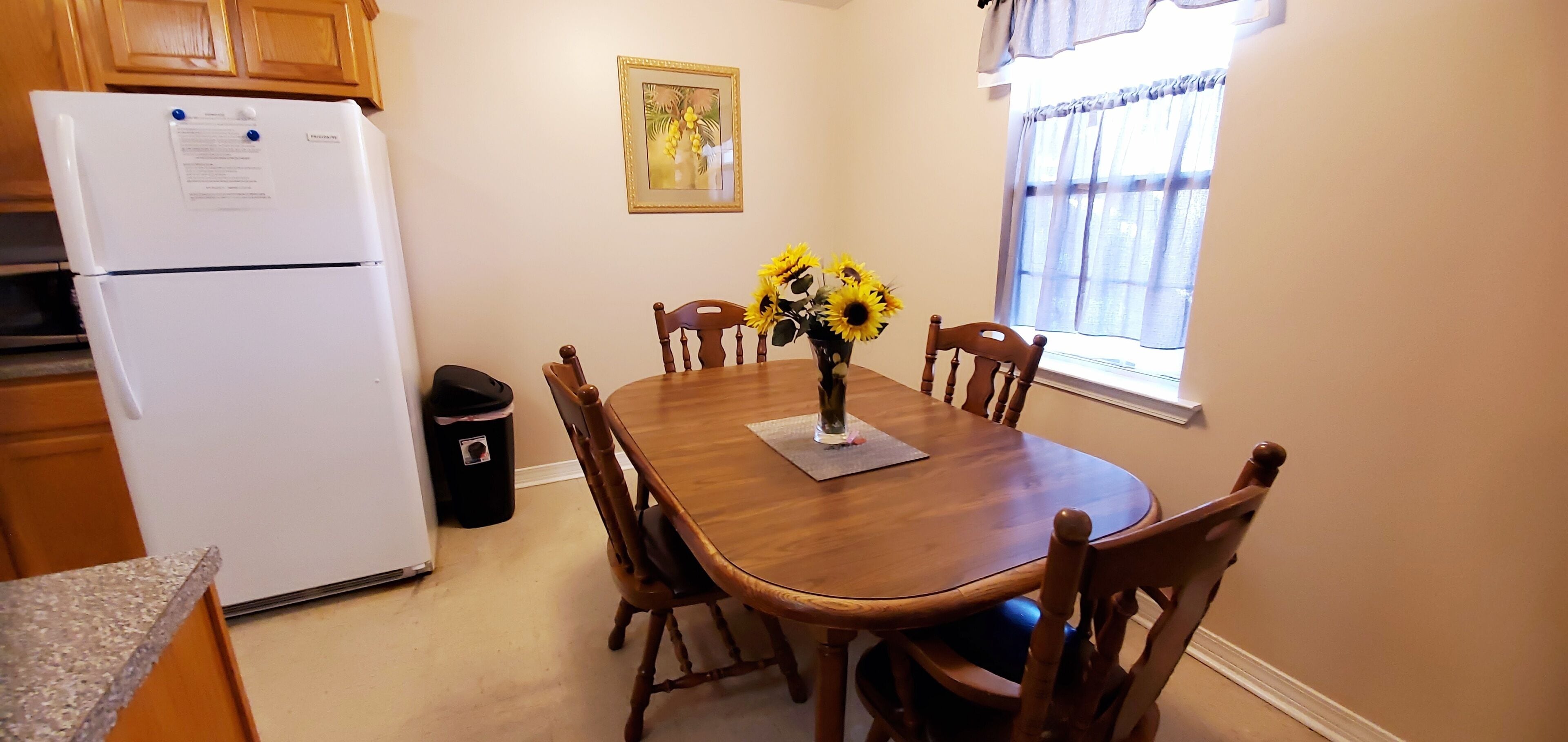 Un coin repas avec une table en bois, six chaises, un vase avec des tournesols, un réfrigérateur, une poubelle et une fenêtre avec des rideaux.