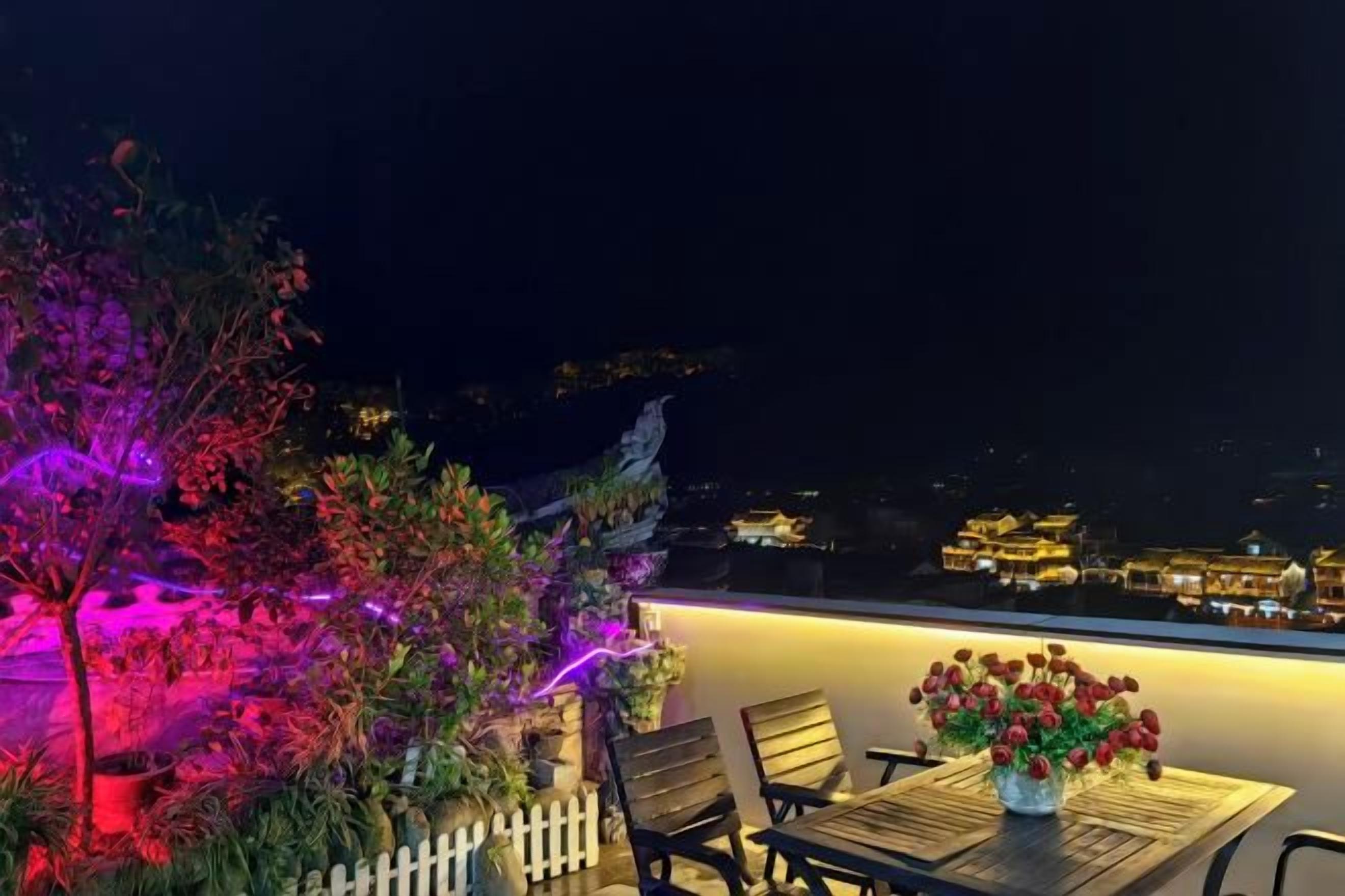 An outdoor seating area with a table and chairs, a vase of flowers, and a view of a cityscape at night.