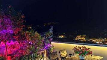 Un área de estar al aire libre con mesa y sillas, un jarrón de flores y vista a un paisaje urbano nocturno.