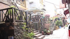 Une ruelle étroite, avec des marches de pierre menant à une porte en bois, bordée d’immeubles dotés de balcons et de plantes en pot.