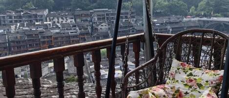 A wicker chair with floral cushions on a balcony overlooking a traditional village with tiered buildings and a bridge.