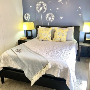 Un letto rifatto con un piumone bianco e cuscini decorati, affiancato da due comodini con lampade, in una stanza con parete scura e decorazioni raffiguranti tarassaci e farfalle.