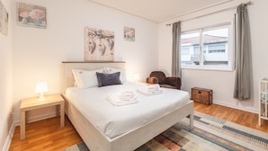 Un dormitorio ordenado con cama, mesita de noche, silla y ventana con cortinas.